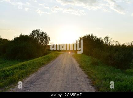 Strada di campagna al tramonto. Strada sterrata con campo agricolo in campagna sullo sfondo di boschi e raggi luminosi del sole Foto Stock