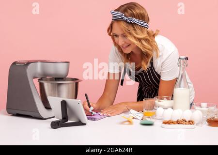 Giovane adulto concentrato femmina prepararsi a fare panetteria, scrivere nuova ricetta da internet, utilizzando tablet digitale, la preparazione di torte. studi interni Foto Stock