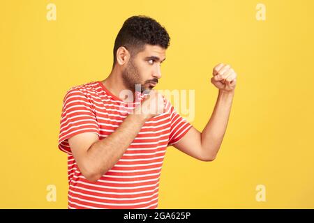 Profilo ritratto concentrato uomo serio con barba in t-shirt a righe che tiene i pugni aggrappati pronti a pugilato, pugnando con espressione sicura. Foto Stock