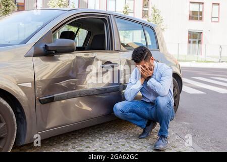 Ritratto di uomo che indossa jeans e camicia blu ha rotto la sua auto, essendo sconvolto e coprendo il suo viso con palme, ammaccature e graffi sulla porta di auto, da Foto Stock