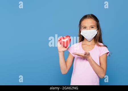 Bambino in maschera medica che punta al cuore decorativo con la mano isolata su blu Foto Stock