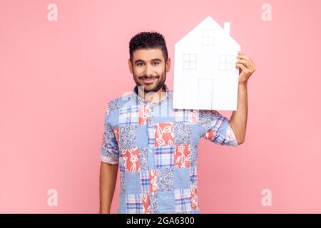 Uomo attraente con barba che indossa una camicia casual blu che tiene la casa di carta guardando la macchina fotografica con un sorriso piacevole, sognando di casa propria. Prigioniero interno Foto Stock