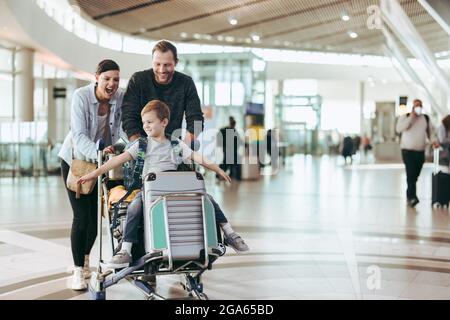 Coppia felice di spingere il trolley con il loro figlio all'aeroporto. I bambini si divertono seduti sul carrello dei bagagli mentre i genitori lo spingono all'aeroporto. Foto Stock