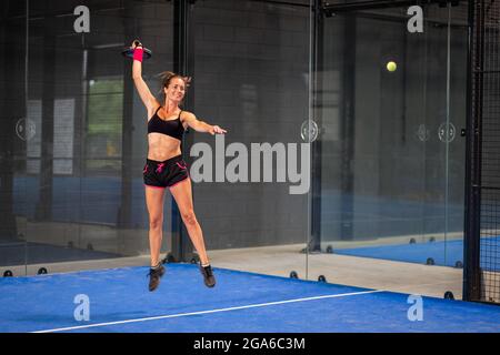 Donna che gioca un padel in un campo di risaie in erba blu indoor - giovane donna sportiva giocatore di risone che colpisce la palla con una racchetta Foto Stock