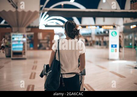 Donna caucasica che cammina attraverso il terminal dell'aeroporto durante la pandemia coviale caricando i suoi bagagli in un trolley facendo tempo fino al suo volo Foto Stock