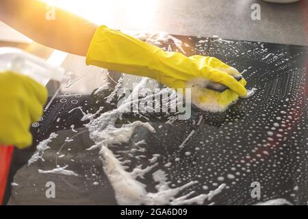 Mano in guanto di gomma per la pulizia stufa elettrica con spruzzatore bottiglia e straccio in una giornata di sole, cucina di pulizia concetto sfondo Foto Stock