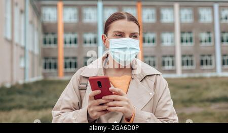 Ritratto di giovane ragazza elegante in maschera medica con moderno smartphone nel parco universitario Foto Stock