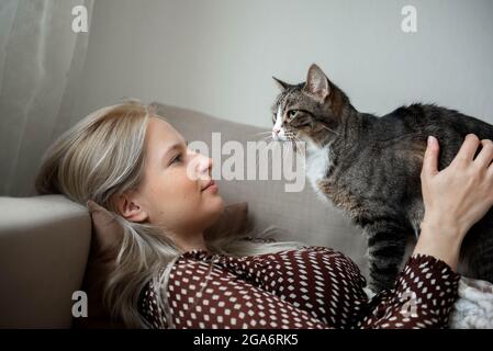 Giovane donna graziosa con gatto domestico cute sta posando sul divano a casa Foto Stock
