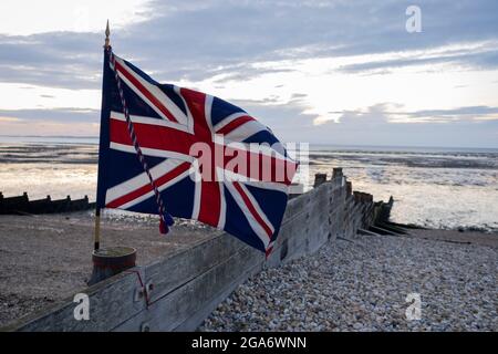 Una bandiera di Union Jack si flutter in una brezza morbida su un tramonto serale di bassa marea sulla spiaggia, il 27 luglio 2021, a Whitstable, Kent, Inghilterra. Foto Stock