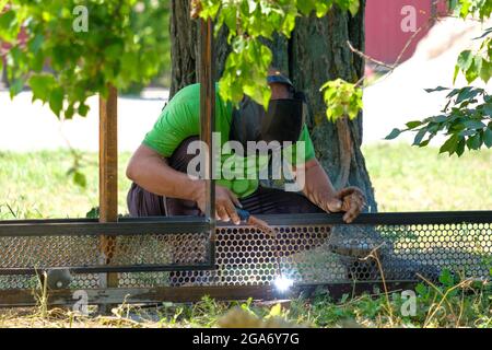 Un operatore in un elmetto di protezione speciale salda il metallo con saldatura ad arco elettrico. Estate giorno di sole. Lampi luminosi e scintille volano ai lati. Foto Stock