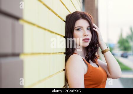 Meravigliosa onda marrone capelli giovane donna playfully posa strada. Foto all'aperto di giovane donna ispirata in maglia arancione che si pone sullo sfondo di un muro di mattoni giallo Foto Stock