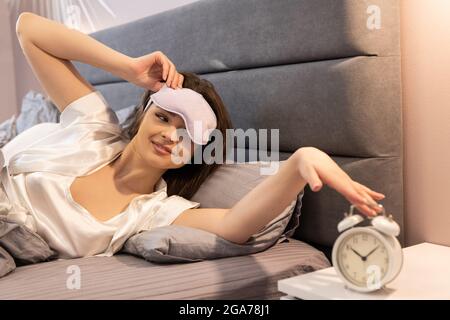 La ragazza ferma la sveglia mentre si sveglia a letto Foto Stock