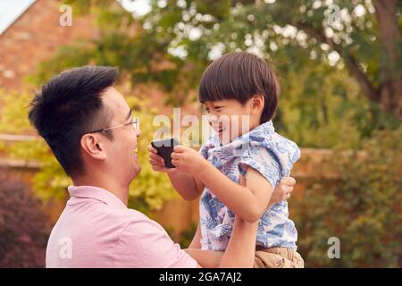 Asian Padre coccolare Figlio in giardino come ragazzo gioca con il cellulare Foto Stock