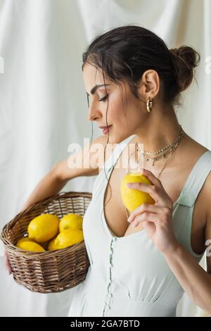 bella donna in abito che tiene il cesto di vimini e limone su bianco Foto Stock