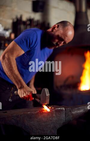Maschio Blacksmith martellando metallo su incudine con Blazing Forge in background Foto Stock