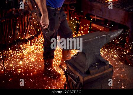 Primo piano di Male Blacksmith Holding Hammer vicino a incudine circondato da Sparks e Embers Foto Stock