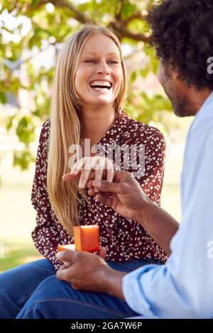 Romantico uomo maturo che propone alla donna sorpresa seduta sul Park Bench con anello di impegno in Box Foto Stock