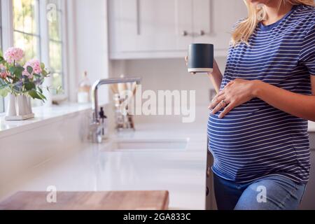 Primo piano di donna incinta in piedi da cucina finestra toccare stomaco rilassante con bevanda calda Foto Stock