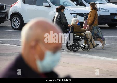 Persone che camminano in una strada con maschera durante i periodi di pandemia. L'operatore sanitario spinge la sedia a rotelle con una donna anziana seduta in essa. Foto Stock