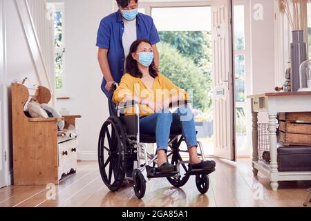 L'uomo asiatico maturo che indossa la maschera di faccia che spinge la moglie in sedia a rotelle a casa durante la Pandemia di salute Foto Stock