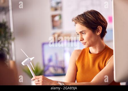 Architetto femminile in ufficio che lavora su disegni per progetto di energia rinnovabile con modello di turbina eolica Foto Stock