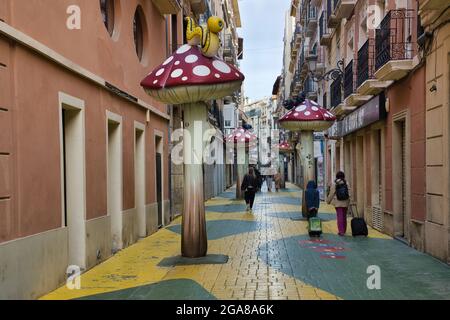 Un vicolo lastricato con alti funghi giganti d'arte distanziato e la gente che cammina nella città di Alicante, Spagna Foto Stock