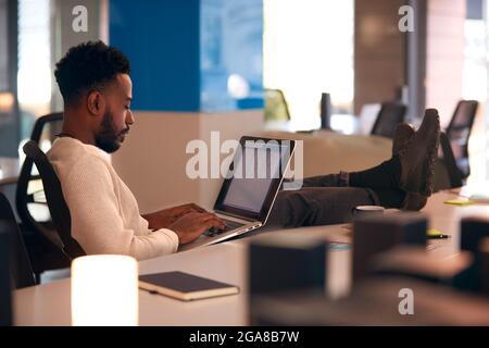 Giovane uomo d'affari che lavora tardi seduto alla scrivania con laptop in un moderno ufficio open space Foto Stock