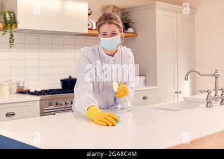 Donna a casa che indossa maschera in cucina facendo casalinghi e pulizia superficie contatore con spray Foto Stock