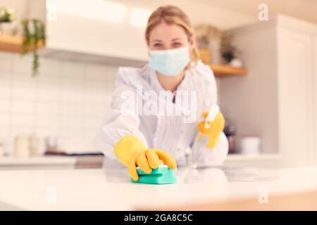 Donna a casa che indossa maschera in cucina facendo casalinghi e pulizia superficie contatore con spray Foto Stock