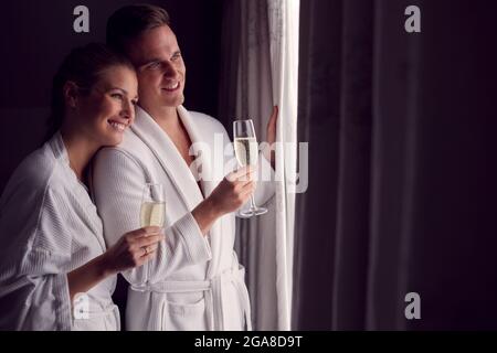 Coppia che indossa accappatoi sul Romantic Hotel Break Standing da tende aperte con bicchieri di Champagne Foto Stock