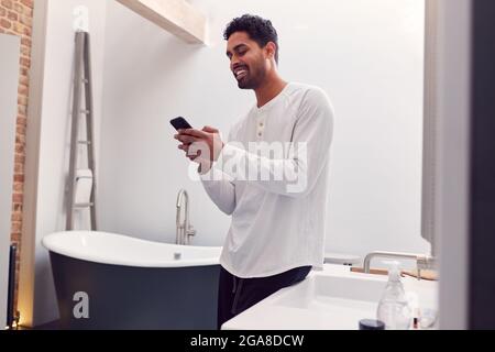 Uomo che indossa Pajamas a casa controllare telefono cellulare in bagno moderno Foto Stock