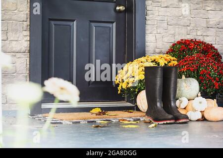 Stivali da pioggia seduti sul tappetino della porta del portico anteriore che è stato decorato per l'autunno con tovaglioli bianchi, arancioni e grigi e le mamme. Foc. Selettivo Foto Stock