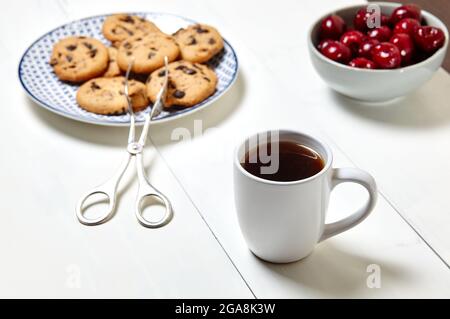 Tazza bianca con caffè, biscotti tradizionali e ciliegie dolci in una ciotola di ceramica su fondo di legno bianco, primo piano. Gustosa colazione, mattina routin Foto Stock