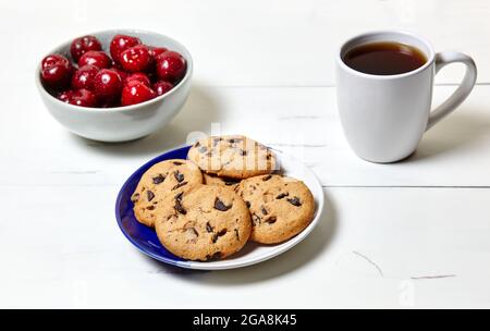 Tazza bianca con caffè, biscotti tradizionali e ciliegie dolci in una ciotola di ceramica su fondo di legno bianco, primo piano. Gustosa colazione, mattina routin Foto Stock
