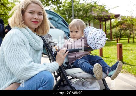 Mamma e figlia sulla natura a piedi presso il parco primaverile. Adorabile bambina in abiti comuni seduta in carrozzina blu. Bambino in buggy Foto Stock