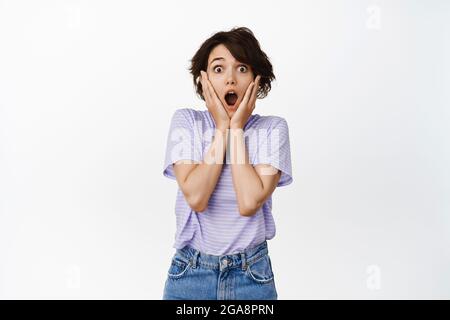 Immagine di sorpresa, bruna ragazza speechless caduta mascella, WOW faccia, tenendo le mani sul viso, fissando impressionato alla macchina fotografica, notizie unbelievable, levandosi in piedi sopra Foto Stock