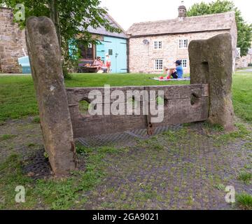 Gli stock di legno nel villaggio della peste del Derbyshire di Eyam. Sullo sfondo ci sono 2 bambini che giocano sul verde racchiuso da 2 edifici in pietra. Foto Stock