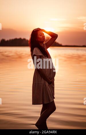 Donna incinta all'aperto. Silhouette di giovane donna felice in raggi di sole che si alza in piedi in acqua sulla riva del lago e che tocca i suoi capelli e stomaco Foto Stock