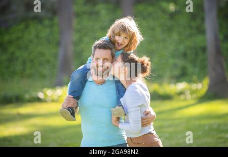 Madre baciare padre mentre bambino ragazzo cavalcare piggy indietro sui padri spalle estate all'aperto, famiglia Foto Stock