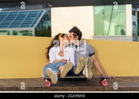Un paio di pattinatori che si baciano si siedono a bordo lungo insieme all'aperto nello spazio urbano. Gioventù, romanticismo e amore Foto Stock