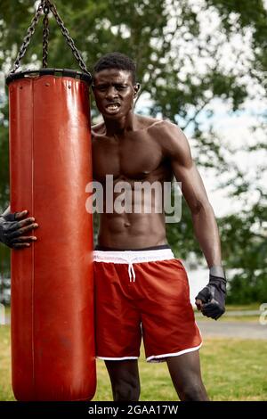 Pugile professionista nero muscolare sudare in guanti di boxe formazione all'aperto, giovane pugile afroamericano che si esercita sul parco giochi di strada. Foto Stock