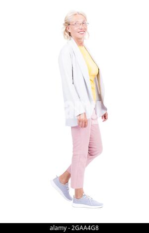 Foto verticale a lunghezza intera di donna caucasica anziana con outfit elegante che cammina, sfondo bianco Foto Stock