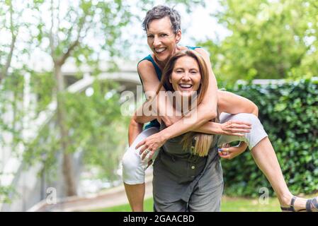 Donna matura che dà il piggyback alla femmina matura amica nel parco Foto Stock