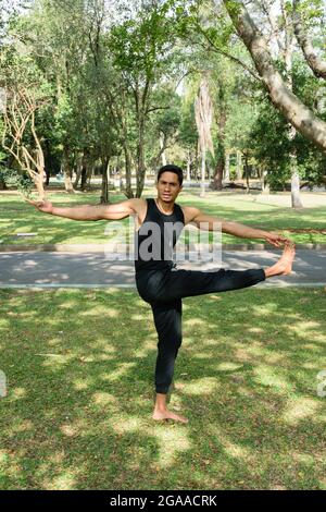 Giovane brasiliano che pratica yoga in una posizione equilibrata in un parco pubblico in una giornata di sole. Foto Stock
