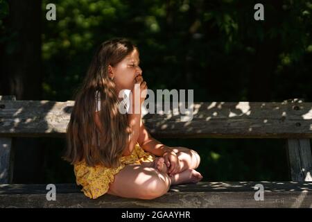 Carino ragazza caucasica divertente seduta sulla panchina e mangiare gelato . Ragazzino sorridente e sognante. Ora legale. Felice infanzia. Foto Stock