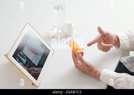 Primo piano del medico seduto al tavolo e puntando alla bottiglia con la medicina mentre si è in contatto online con il suo collega su tablet digitale Foto Stock
