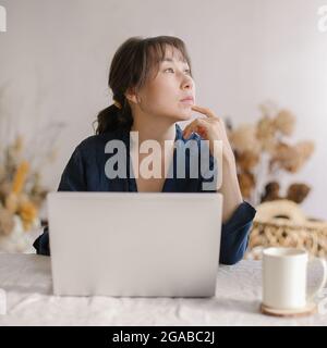 Pensieroso giovane donna asiatica d'affari guardando da parte e pensando a qualcosa mentre lavora da casa Foto Stock
