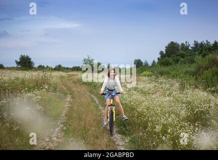 divertente ritratto di bella ragazza emotiva 17-18 anni su una bicicletta in movimento. divertirsi a cavallo di una bicicletta nei pittoreschi campi fioriti. sano Foto Stock