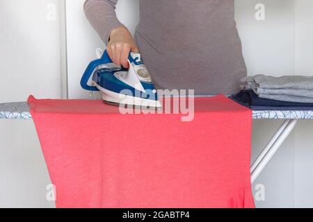 Casalinghi e concetto di casa. La donna stira la T-shirt corallina a casa. La mano femminile sta stirando i vestiti, primo piano. Foto Stock