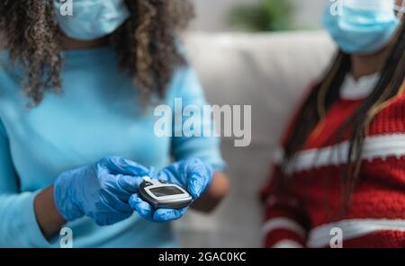 Giovane infermiera che controlla il test glicemico per il diabete al paziente anziano mentre si indossano guanti chirurgici e maschera facciale - concetto sanitario Foto Stock